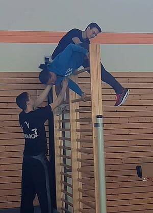 Kind klettert über Sprossenwand und zwei Personen helfen