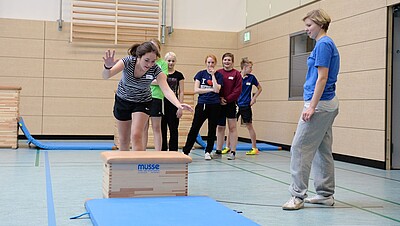 Übungsleitende im Sportunterricht leitet eine Sprung über kleinen Kasten an.