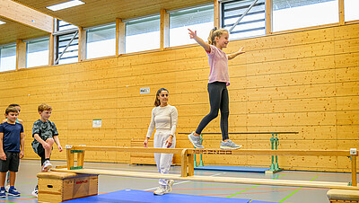 Mädchen balanciert vor Lehrerin auf Balken.