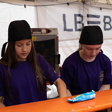 Symbolbild zwei Teilnehmerinnen bei der Ausgabe von HotDogs beim Tag des offenen Zeltes im Camp Breisach 2026 